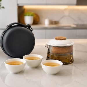 May include: A glass teapot with a wooden collar and white lid, three white ceramic tea cups filled with light amber tea, and a round black carrying case with a handle and zip closure, arranged on a white marble countertop surface.