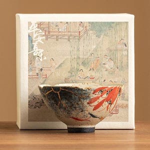 May include: A ceramic bowl with a unique design, featuring a blend of blue, gold, and red hues. The bowl is set against a backdrop of a box with an Asian-inspired illustration. The bowl's design includes a fish motif.
