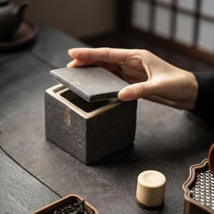 Peut inclure: Un coffret à thé carré en céramique gris foncé avec un couvercle soulevé par une main. Le coffret a une surface texturée et un intérieur de couleur claire. Un petit cylindre en bois et un plateau de feuilles de thé sont également visibles.