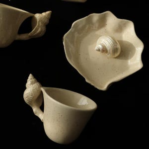 May include: Cream-coloured ceramic teacups and a small plate with a seashell design. The teacups have handles shaped like seashells. The plate has a seashell in the centre. The items are speckled and have a rustic appearance.