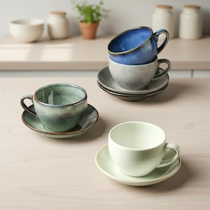 Puede incluir: Una colección de tazas de té y platillos de cerámica en varios colores, incluyendo azul, verde y verde claro. Las tazas están apiladas y dispuestas sobre una superficie de color claro, sugiriendo una cocina o comedor.