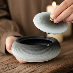 May include: A ceramic ashtray with a lid. The ashtray is round with a black interior and a gradient exterior, transitioning from black to gray. The lid has a gold-colored accent. The ashtray is on a wooden surface.