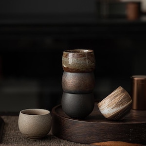 Può includere: Quattro tazze da tè in ceramica fatte a mano in varie tonalità di terra: marrone, bronzo e nero. Una tazza è inclinata su un vassoio di legno, mostrando un motivo a strisce. Le tazze sono progettate per tè o caffè.