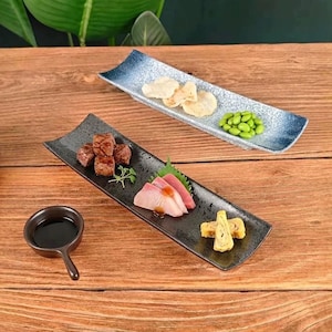 May include: Two rectangular ceramic serving plates with curved edges and a speckled finish. One plate is black, holding cubed meat, sashimi, and rolled egg. The other is blue and white, holding chips and edamame. A small black sauce bowl is included.