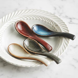 May include: Four ceramic soup spoons in shades of red, blue, gray, and cream rest on a white, textured plate. The spoons have a curved design and are arranged in a fan shape. The plate has a decorative, leaf-like pattern around the rim.