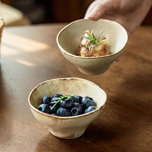 Op de afbeelding: Twee kleine, beige keramische kommen. Eén kom is gevuld met bosbessen en een takje groen. De andere kom, vastgehouden door een hand, bevat een kleine portie voedsel gegarneerd met rozemarijn. De kommen hebben een rustieke, handgemaakte uitstraling.
