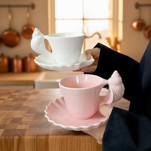 May include: Two ceramic teacups and saucers. One is white, the other is pink. Both have a seashell-shaped handle and a scalloped edge on the saucer. The teacups are being held in front of a kitchen background.