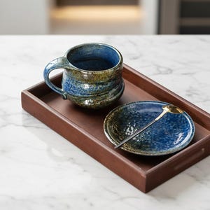 Könnte beinhalten: Ein handgefertigtes Keramik-Tassen- und Untertassen-Set mit einem goldfarbenen Löffel, präsentiert auf einem Holztablett. Die Tasse und die Untertasse haben eine blaue und grüne Glasur. Der Löffel hat einen schwarzen Griff. Das Tablett ist dunkelbraun.