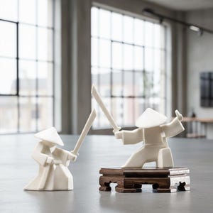 May include: Two white ceramic samurai figurines in a fighting pose, each holding a sword. One figurine is kneeling on a small, dark wooden stand. The figures wear traditional conical hats and robes. The background is a neutral-toned room with large windows.
