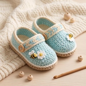May include: A pair of light blue crochet slippers with a cream-colored trim and accents. Each slipper features a wooden button, a daisy, and a bee applique. The slippers are displayed on a wooden surface with a crochet hook and wooden beads.