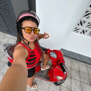 Op de afbeelding: Een felrode motorscooter met een bruin zadel en een zilverkleurige voetenplank. De bestuurder draagt een roze helm met een doorzichtig vizier, een zonnebril met een zwart montuur en gele glazen, en een rood-zwart mouwloos topje met uitsnijdingen.