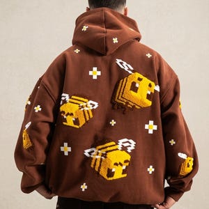 May include: A brown hooded sweatshirt featuring pixelated yellow and black bees and white cross accents. The back of the sweatshirt displays multiple embroidered bees and crosses, creating a unique and playful design. The hoodie has a drawstring and a kangaroo pocket.
