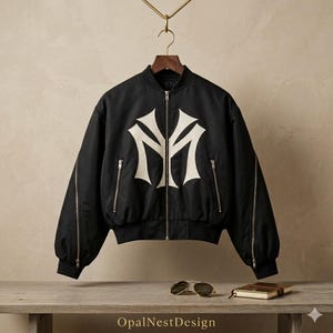 Könnte beinhalten: Schwarze Bomberjacke mit weißem Logo auf der Vorderseite. Die Jacke hat einen Reißverschluss, zwei Seitentaschen sowie gerippte Bündchen und Saum. Sie hängt an einem Holzkleiderbügel.