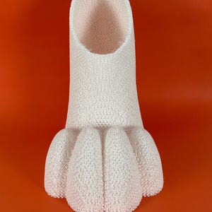 May include: A white, textured, cylindrical object with a hollow top opening and a base featuring five rounded, protruding, paw-like segments. The material has a porous, mesh-like appearance throughout the entire structure of the item.