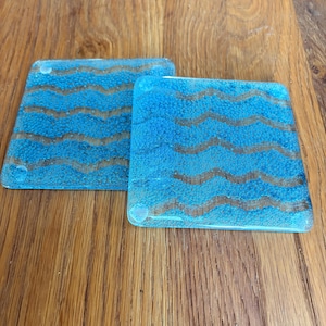 May include: Two square glass coasters with a blue, textured surface and wavy brown lines. The coasters are placed on a wooden surface. The coasters are a decorative item.