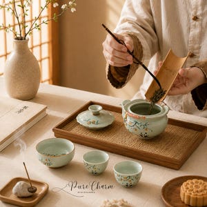 May include: A light green ceramic tea set featuring hand-painted panda and peach branch motifs, including a lidded teapot, three small cups, and a wooden serving tray. A bamboo scoop is used to pour loose tea leaves into the teapot.
