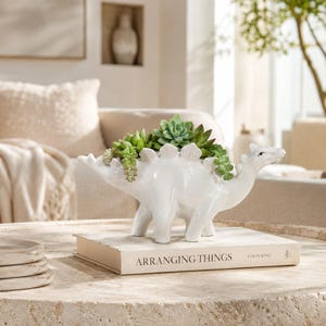 May include: A white ceramic dinosaur-shaped planter featuring a row of plates along its back, filled with green succulent plants. The planter is positioned on top of a white book with the text Arranging Things by Colin King printed on the cover.