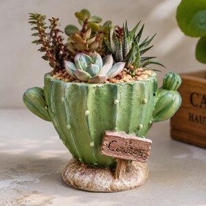 Puede incluir: Una maceta de cerámica verde con forma de cactus, dos brazos laterales y superficie texturizada. Un pequeño letrero de estilo madera fijado en la parte frontal tiene escrita la palabra "Cactus". La maceta contiene varias suculentas pequeñas.