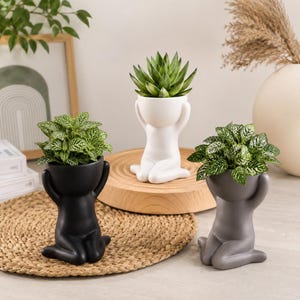 Puede incluir: Tres macetas de cerámica con forma de figuras arrodilladas con los brazos levantados sobre la cabeza. Una maceta es negra mate, otra blanca mate y otra gris mate. Cada figura sostiene una pequeña planta verde con hojas variegadas.