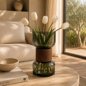 Può includere: Un vaso decorativo composto da due sezioni in vetro grigio scuro sovrapposte, separate da una fascia centrale cilindrica in legno, contenente cinque tulipani bianchi con steli verdi, posizionato su un tavolo rotondo in pietra.