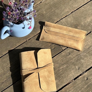 May include: A light brown suede journal with a wrap-around tie closure and a matching rectangular suede envelope pouch, positioned near a light blue ceramic teapot containing a bouquet of dried purple and pink flowers.