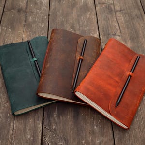 Pode incluir: Três diários encadernados em couro nas cores verde-escuro, castanho-escuro e castanho-avermelhado. Cada diário apresenta um design de capa envolvente com uma presilha dedicada que segura uma caneta preta fina. Os diários estão dispostos lado a lado, exibindo as suas capas de textura mate e lisa e as páginas de papel branco visíveis ao longo das margens.
