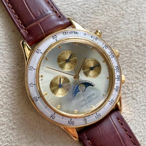May include: Gold-toned wristwatch with a white bezel and a brown leather strap. The watch face features multiple subdials, a moon phase indicator, and the text "TACHYMETER" and "HELBROS".