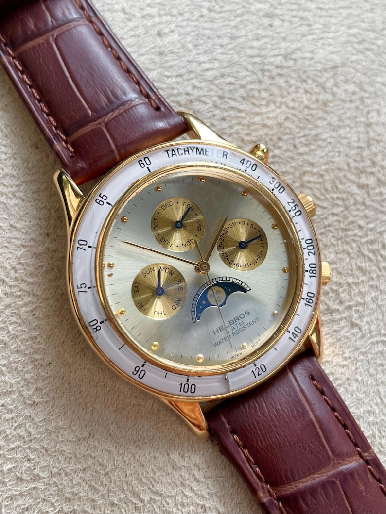 May include: Gold-toned wristwatch with a white bezel and a brown leather strap. The watch face features multiple subdials, a moon phase indicator, and the text "TACHYMETER" and "HELBROS".