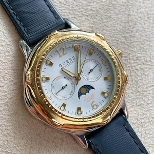 May include: A Guess Waterpro watch with a gold-tone bezel and a white face. The watch features a black leather strap and subdials for the day and date. The watch is water-resistant up to 50 metres.