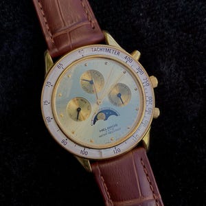 May include: Gold-toned Helbros watch with a light blue face, featuring multiple subdials and a moon phase indicator. The watch has a brown leather band and a tachymeter bezel. The watch is water resistant.