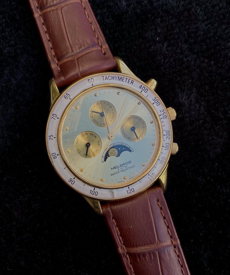 May include: Gold-toned Helbros watch with a light blue face, featuring multiple subdials and a moon phase indicator. The watch has a brown leather band and a tachymeter bezel. The watch is water resistant.