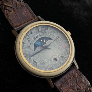 Könnte beinhalten: Fossil Armbanduhr mit Mondphasenzifferblatt und braunem Lederarmband. Das Zifferblatt zeigt ein detailliertes Mondoberflächendesign mit einer amerikanischen Flagge und dem Fossil-Logo. Das Armband ist geflochten.