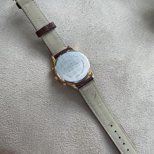 May include: Gold-toned wristwatch with a brown leather band. The watch has a round face and a stainless steel back. The band is light beige on the inside and dark brown on the outside. The watch band measures approximately 9 inches in length.