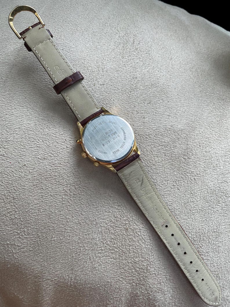 May include: Gold-toned wristwatch with a brown leather band. The watch has a round face and a stainless steel back. The band is light beige on the inside and dark brown on the outside. The watch band measures approximately 9 inches in length.