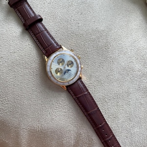 May include: A gold-toned wristwatch with a brown leather band. The watch face is white with gold accents and three smaller dials. The band has a crocodile pattern. The watch is a classic accessory.