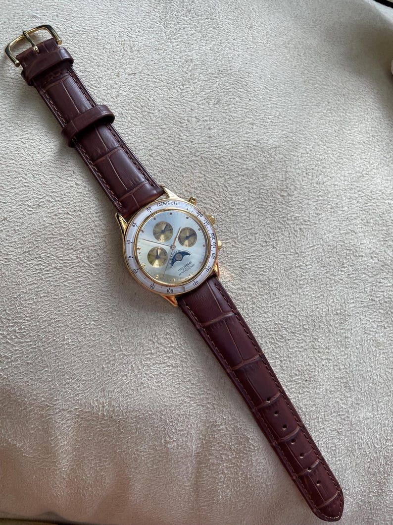 May include: A gold-toned wristwatch with a brown leather band. The watch face is white with gold accents and three smaller dials. The band has a crocodile pattern. The watch is a classic accessory.