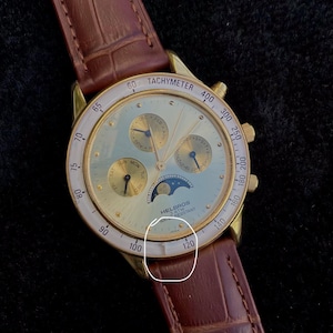 May include: A gold-toned Helbros wristwatch with a brown leather band. The watch face has multiple subdials, a moon phase indicator, and a tachymeter scale. The watch is water resistant and has the text "Japan" on the face.
