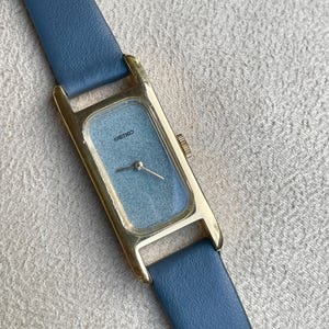 May include: A rectangular gold-toned wristwatch with a light blue face and a blue leather band. The watch face has the brand name "SEIKO" printed on it. The watch band is a solid blue color.