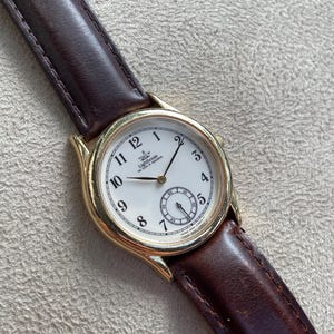 Op de afbeelding: Een goudkleurig polshorloge met een witte wijzerplaat en zwarte cijfers. Het horloge heeft een kleine secondenwijzer en een bruine leren band. De wijzerplaat toont de tekst "Lighthouse" en "Swiss Made". De band is ongeveer 20 cm lang.
