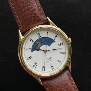 May include: An Alba Quartz wristwatch with a gold-tone round case and a textured brown leather strap. The white dial features black Roman numeral hour markers, thin black hands, and a blue moon phase sub-dial with a yellow crescent moon and stars.
