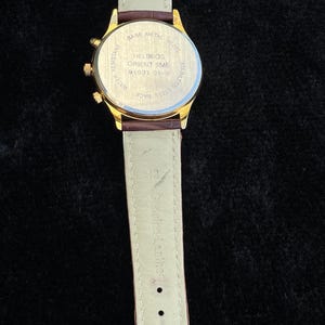 May include: A gold-toned Helbros Orient 9ME watch with a brown leather strap. The watch has a cream-colored band and a gold buckle. The watch face is visible, with text that reads "HELBROS ORIENT 9ME 91931 91-9".