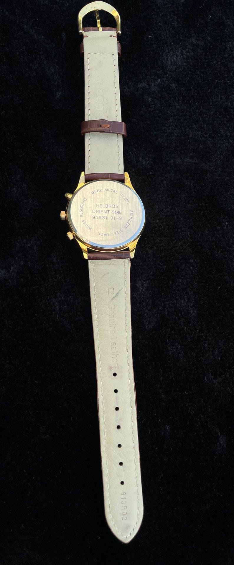 May include: A gold-toned Helbros Orient 9ME watch with a brown leather strap. The watch has a cream-colored band and a gold buckle. The watch face is visible, with text that reads "HELBROS ORIENT 9ME 91931 91-9".