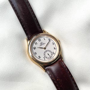 May include: A gold-tone wristwatch with a white dial, black Arabic numerals, and a dark brown leather strap. The dial features the text Lighthouse The Pride of Tomorrow and a small sub-dial for seconds. The watch has thin gold-tone hands.