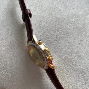 May include: A gold-toned wristwatch with a brown leather band. The watch face has a light-colored dial with small markings and a gold bezel. The band is a deep brown color.