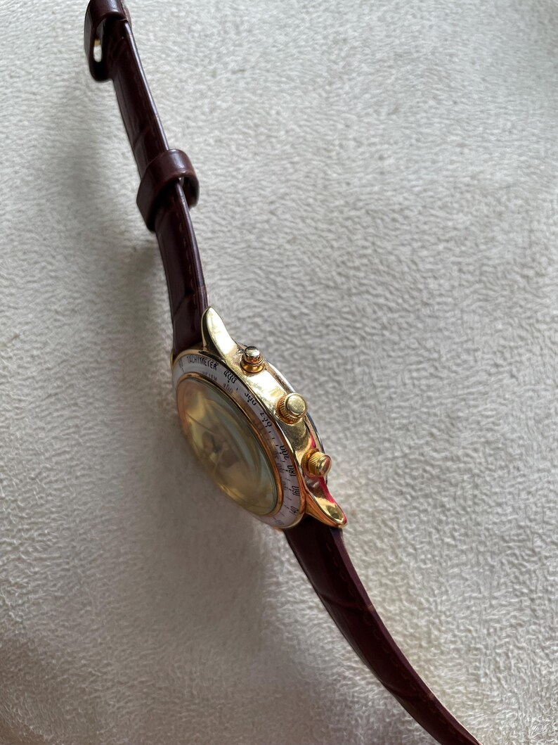 May include: A gold-toned wristwatch with a brown leather band. The watch face has a light-colored dial with small markings and a gold bezel. The band is a deep brown color.