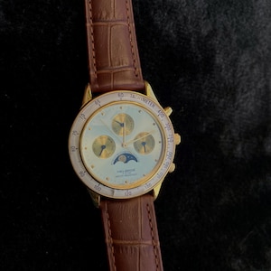 May include: A gold-toned Helbros watch with a light-colored face, featuring multiple subdials and a tachymeter scale. The watch has a brown leather strap with a crocodile pattern. The watch face displays the text "HELGROS" and "WATER RESISTANT".