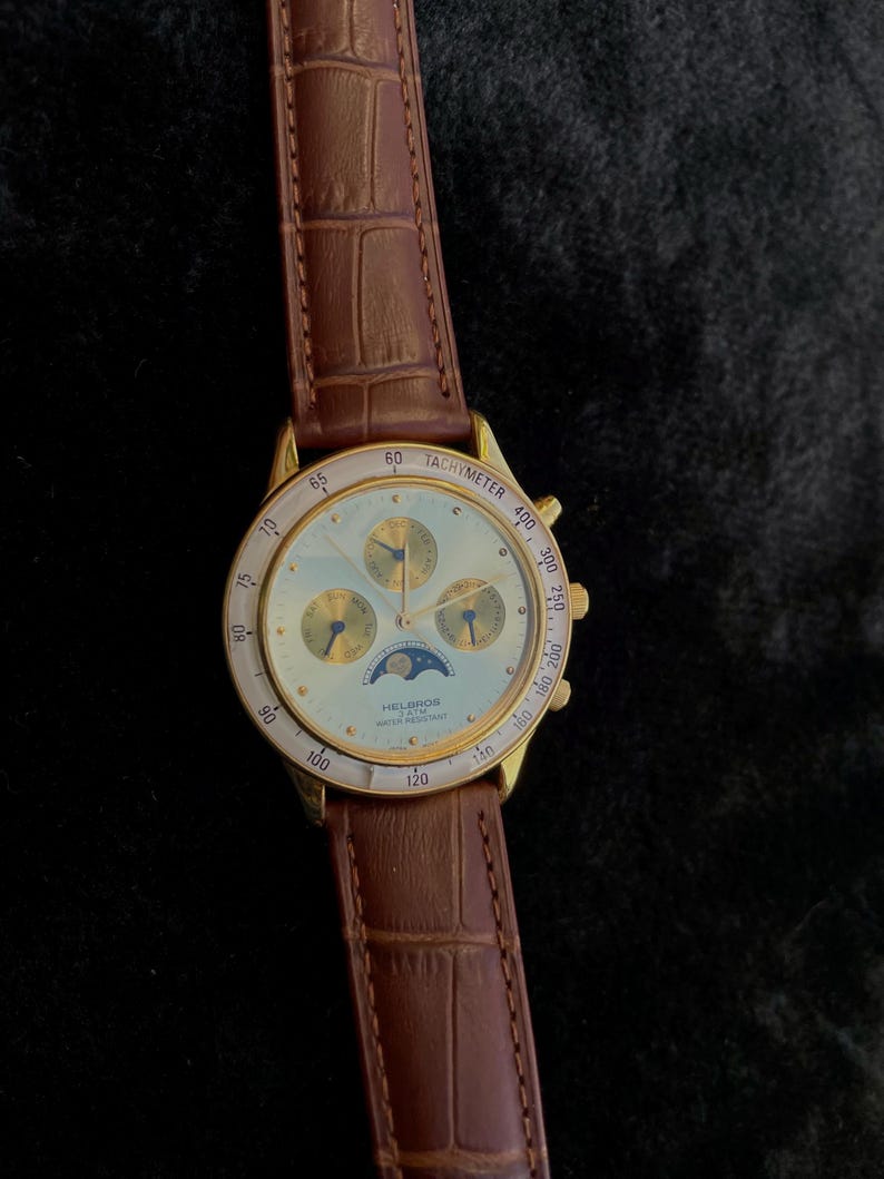May include: A gold-toned Helbros watch with a light-colored face, featuring multiple subdials and a tachymeter scale. The watch has a brown leather strap with a crocodile pattern. The watch face displays the text "HELGROS" and "WATER RESISTANT".