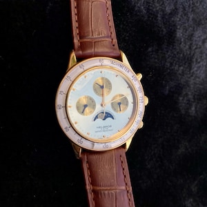 May include: A gold-toned Helbros watch with a white face, featuring subdials and a moon phase indicator. The watch has a tachymeter bezel and a brown leather strap. The watch face displays the text "HELBROS WATER RESISTANT".