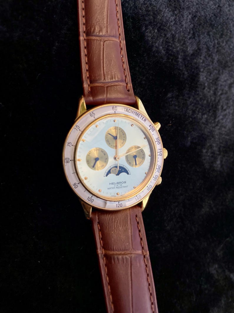 May include: A gold-toned Helbros watch with a white face, featuring subdials and a moon phase indicator. The watch has a tachymeter bezel and a brown leather strap. The watch face displays the text "HELBROS WATER RESISTANT".