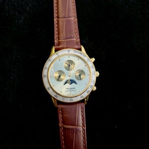 May include: Gold-toned Helbros watch with a light-colored face, featuring multiple subdials and a moon phase indicator. The watch has a brown leather band with a crocodile pattern. The watch face displays the text "TACHYMETER" and "WATER RESISTANT".